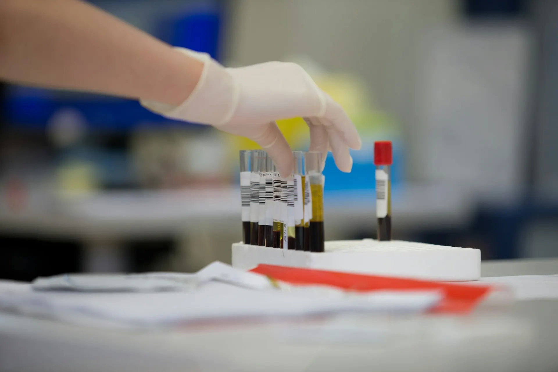 A gloved hand picking up a blood sample tube from a rack in a laboratory